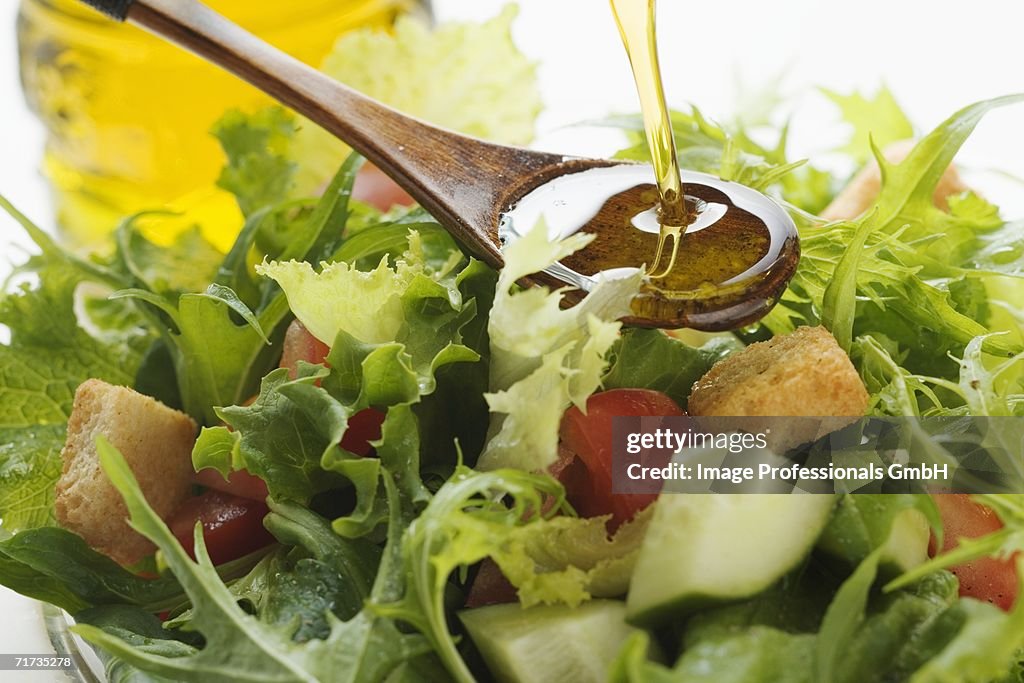 Pouring olive oil into wooden spoon above salad leaves