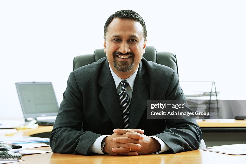 Businessman at desk, smiling at camera, hands clasped