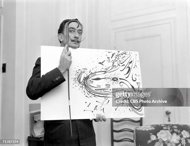 Spanish surrealist painter Salvador Dali holds up a painting and a cane for a broadcast of the CBS television program 'Person to Person' hosted by...