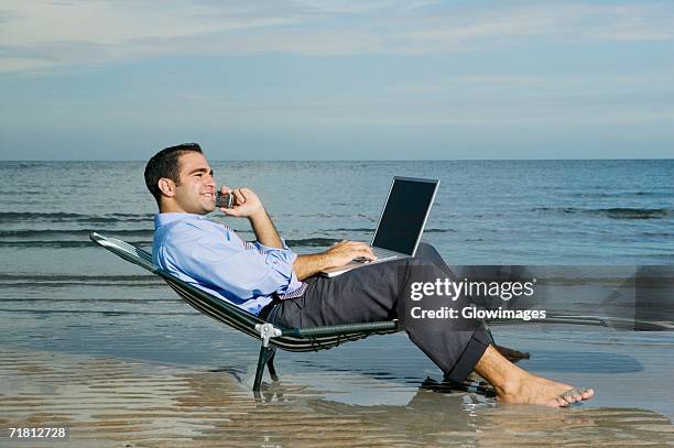 side profile of a young man sitting on a lounge chair on the beach talking on a mobile phone and using a laptop - profile laptop sitting stock pictures, royalty-free photos & images