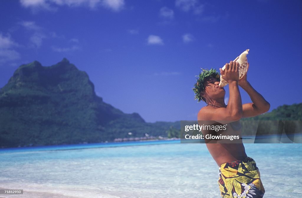 Side profile of a young man blowing a conch shell on the beach, Hawaii, USA