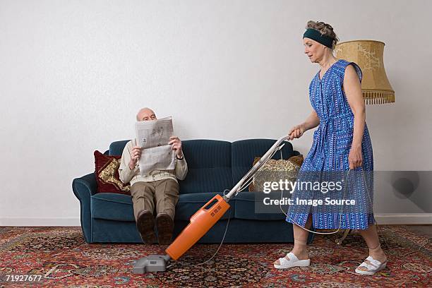 man reading newspaper as woman vacuums - casalinga foto e immagini stock
