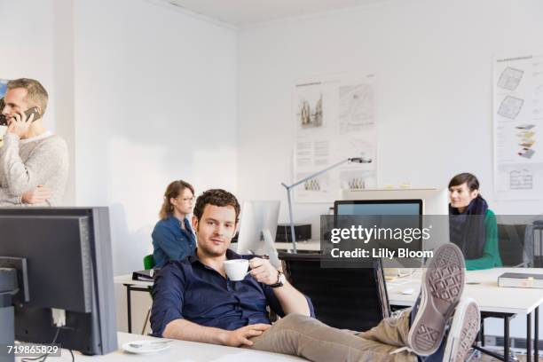 mid adult man in office, feet on desk holding teacup looking at camera - man feet up on desk stock pictures, royalty-free photos & images
