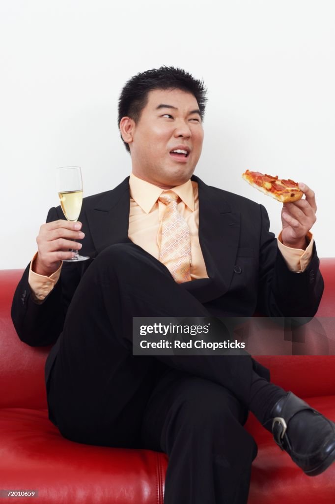 Close-up of a businessman sitting on a couch holding a slice of pizza with a champagne flute