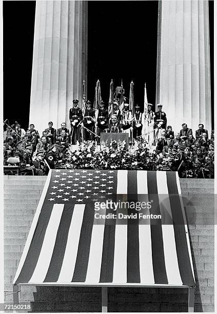 American evangelist Billy Graham spreads his arms in a wide gesture as he speaks to a crowd from a podium above a giant American flag on the steps of...