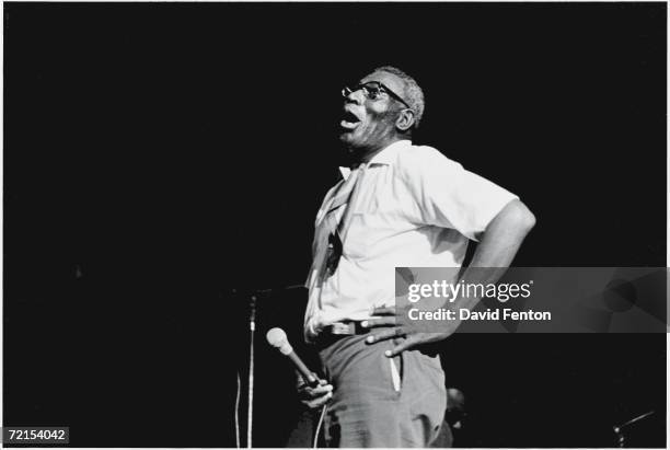 American blues singer and musician Howlin' Wolf holds a microphone and stands on a darkened stage with his hand on his hip as he performs at the...