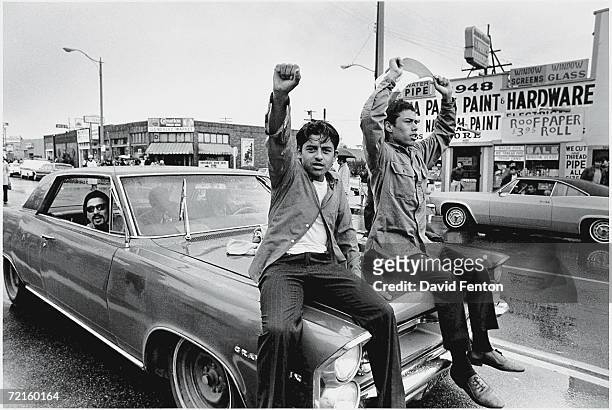 Two young Chicano men ride on the hood of a car and raise their fist during a National Chicano Moratorium Committee march in opposition to the war in...
