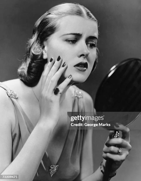 1950s: Portrait of upset woman looking in hand mirror.