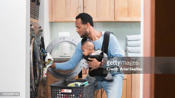 black father with son in baby carrier doing laundry - portabebés fotografías e imágenes de stock