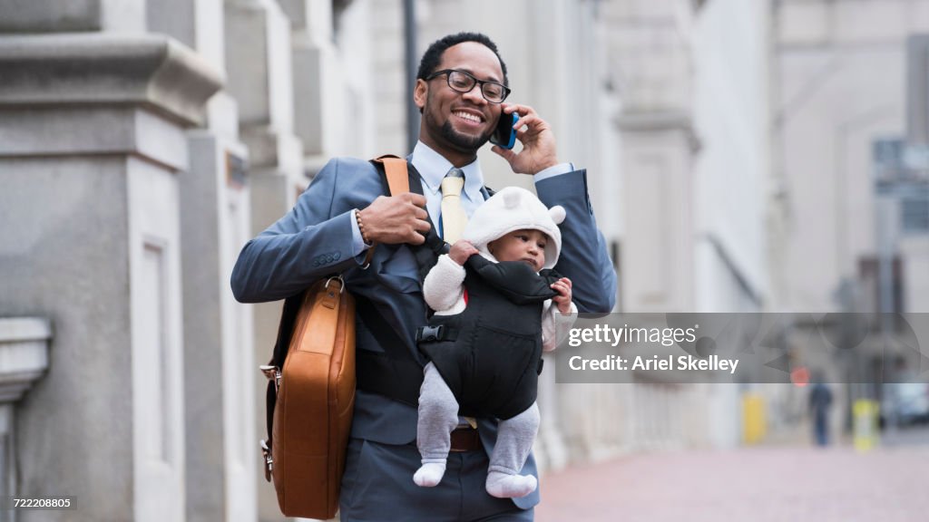 Black businessman with son in baby carrier talking on cell phone