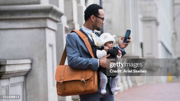 black businessman with son in baby carrier texting on cell phone - portabebés fotografías e imágenes de stock