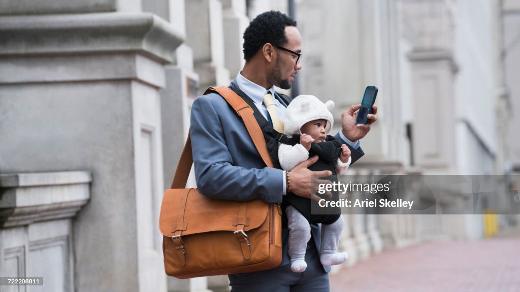 Black businessman with son in baby carrier texting on cell phone