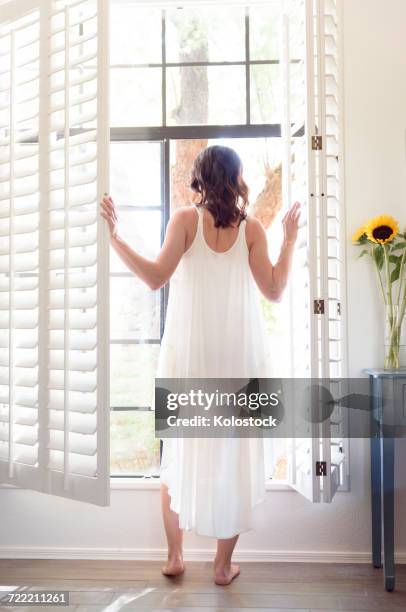 caucasian woman wearing nightgown opening window shutters - negligée stock-fotos und bilder