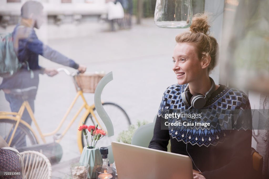 Smiling young woman with headphones using laptop in urban cafe window