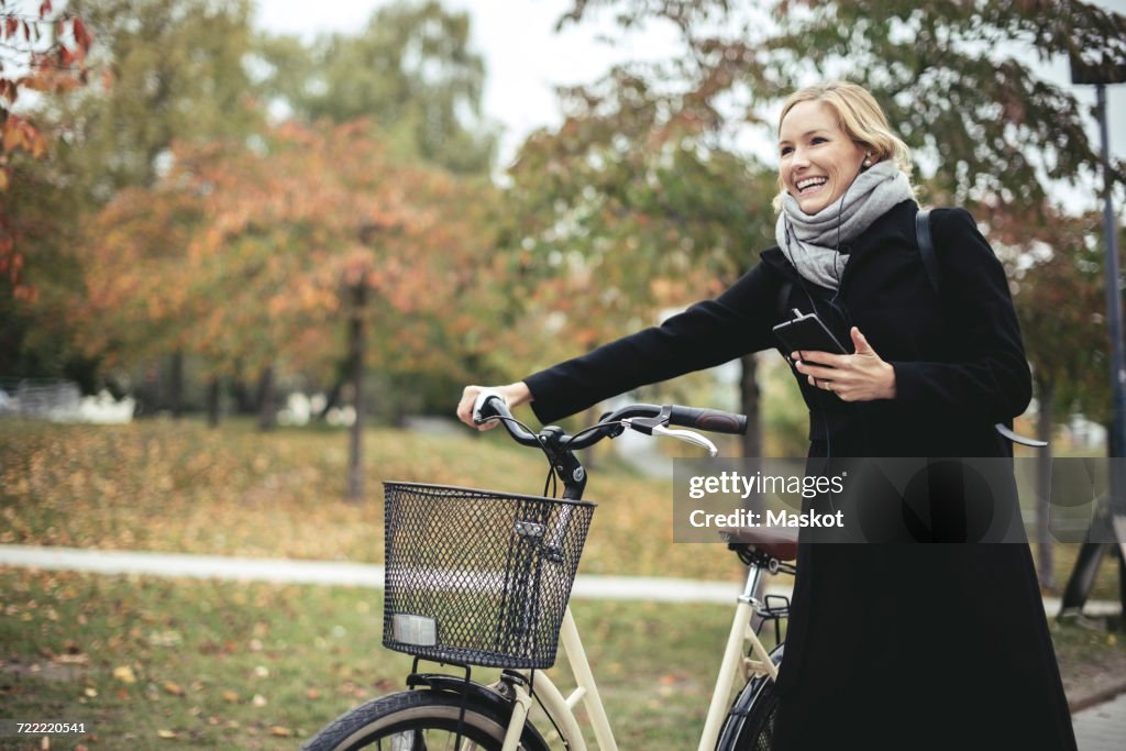 Smiling businesswoman enjoying music while walking with bicycle during autumn