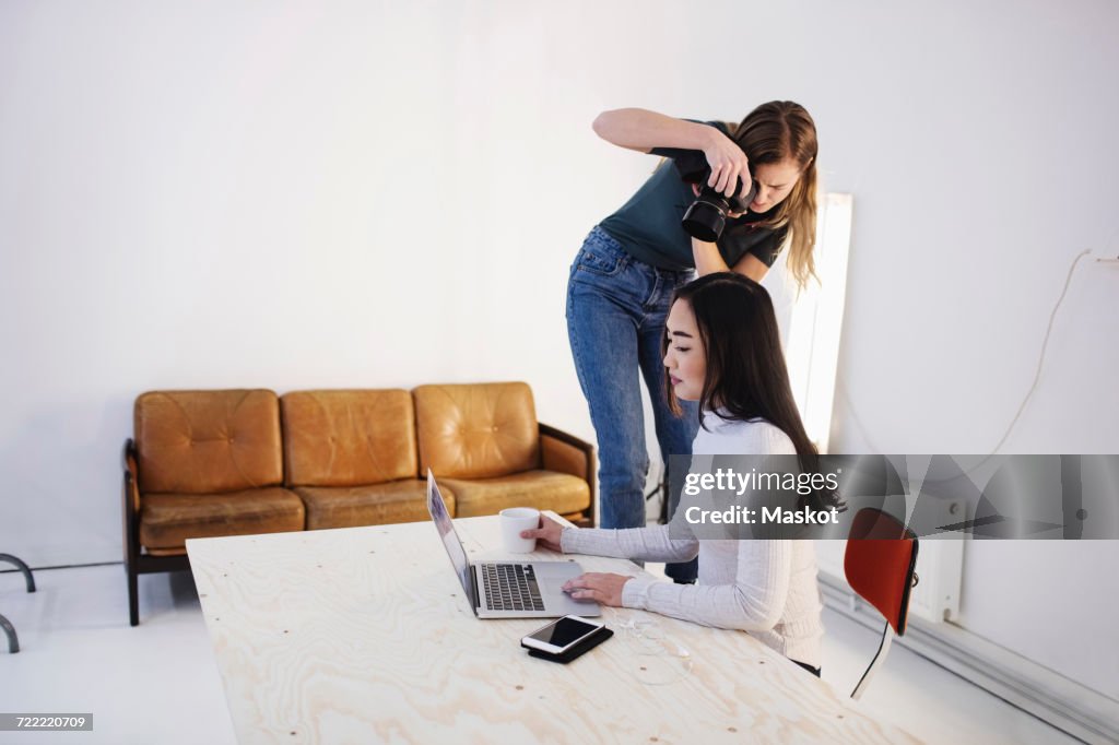 Female blogger photographing colleague using laptop at desk in creative office