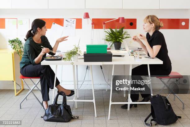 full length of businesswomen talking while having lunch at table in office - lunch argument stock-fotos und bilder