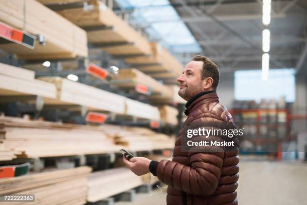 customer looking at wooden planks on shelves while holding smart phone in hardware store warehouse - bouwmarkt stockfoto's en -beelden