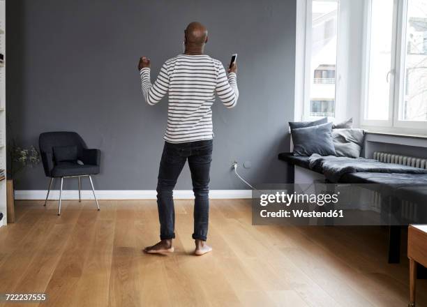 mature man dancing alone at home, holding smart phone - barefoot men stock-fotos und bilder