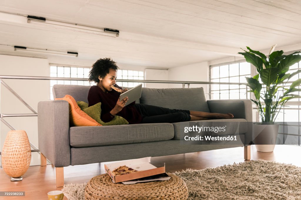 Young woman lying on couch using tablet and eating pizza