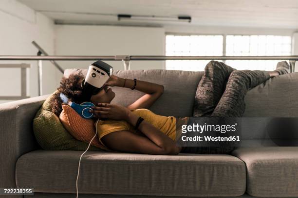 young woman lying on couch with headphones and vr glasses - casques réalité virtuelle photos et images de collection