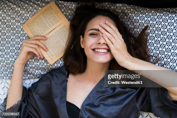 portrait of smiling young woman lying on bed with a book covering one eye with her hand - beautiful women bed stockfoto's en -beelden