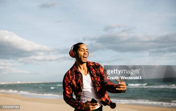 portrait of young man on the beach singg and dancing while listening music with headphones - man singing stockfoto's en -beelden
