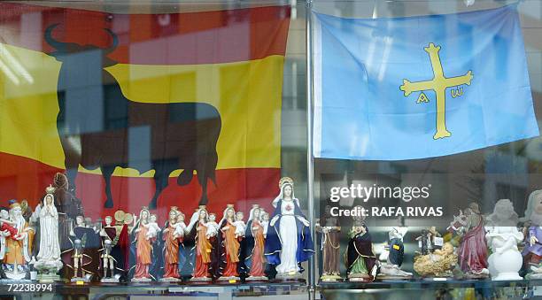 Porcelain figurines of the virgin Mary and Catholic saints with a backdrop of Spanish and Asturias flags on show in a shop window in the square where...