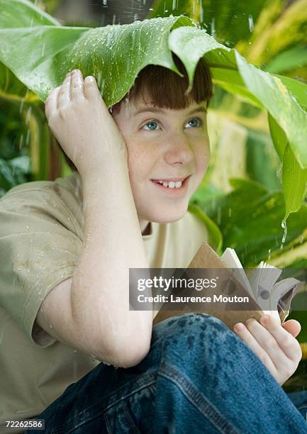boy with book sitting under leaf in rain - makeshift shelter stock pictures, royalty-free photos & images