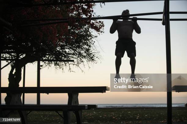 full length of man doing chin-ups at park against sky at sunset - chin ups stock pictures, royalty-free photos & images