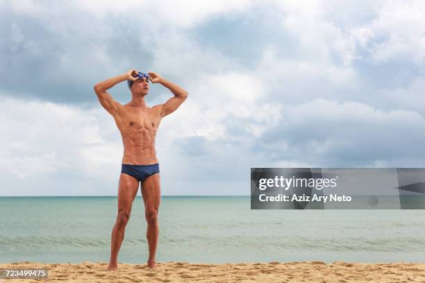 muscular male swimmer standing on beach putting on swimming goggles - muscle men at beach stock-fotos und bilder