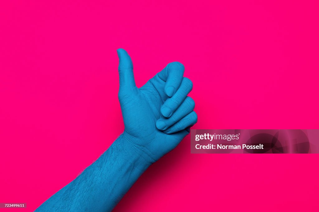 Close-up of blue painted hand gesturing thumb up against pink background