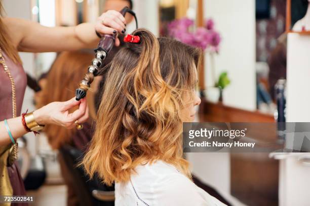 hairdresser using curling tongs on customers long brown hair in salon - fer à friser photos et images de collection