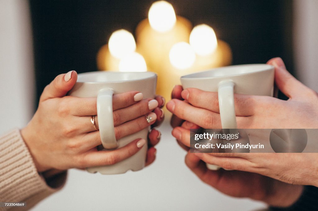 Cropped image of couple holding coffee cups against illuminated candles at home