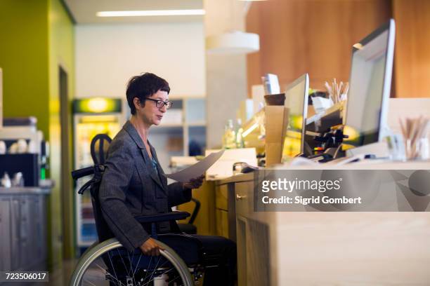 Woman in wheelchair, sitting at desk, looking at document