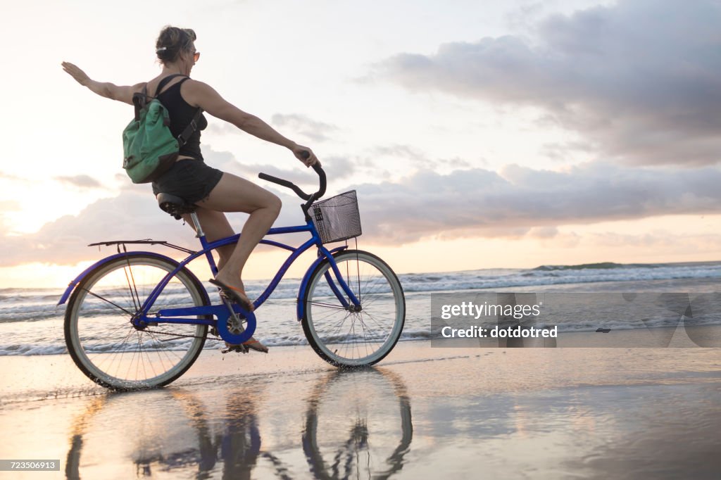 Woman waving while cycling on beach at sunset, Nosara, Guanacaste Province, Costa Rica
