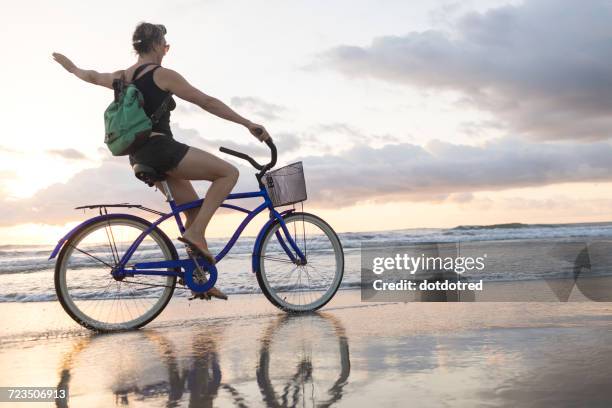 woman waving while cycling on beach at sunset, nosara, guanacaste province, costa rica - nosara costa rica stock pictures, royalty-free photos & images