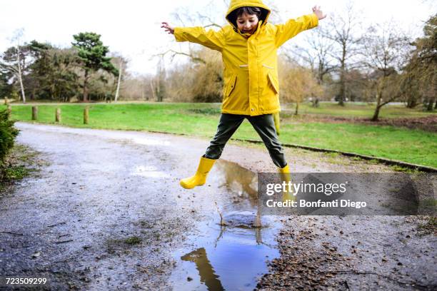 boy in yellow anorak jumping above puddle in park - puddle stock pictures, royalty-free photos & images