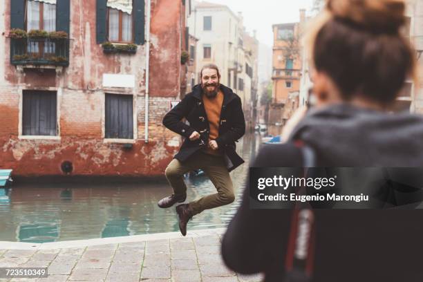 woman photographing man jumping by canal, venice, italy - venice italy stock-fotos und bilder