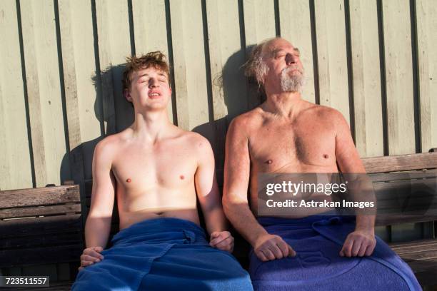 Father and son resting on bench with closed eyes against wooden wall