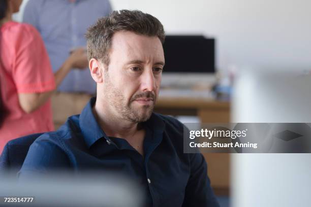 man working at computer on office desk, colleagues on coffee break in background - navy blue interior stock pictures, royalty-free photos & images