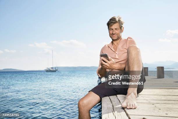smiling man sitting on jetty using cell phone - male knee stock pictures, royalty-free photos & images