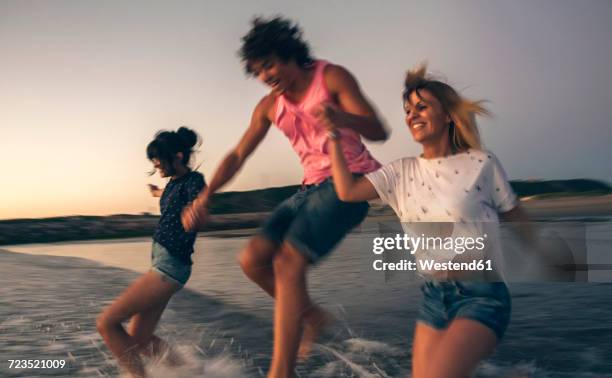 happy friends holding hands and running into the sea at dusk - running shorts foto e immagini stock