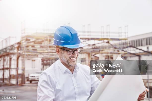 portrait of laughing man wearing blue hart hat looking at construction plan - hart building stock pictures, royalty-free photos & images