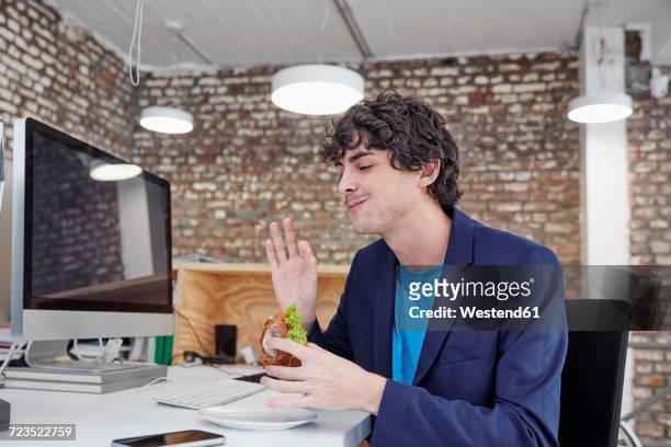 young man sitting at desk, eating sandwich - indulgence photos et images de collection