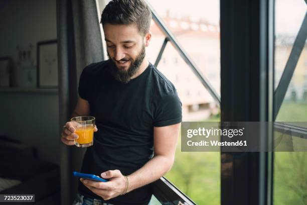 young man drinking an orange juice and using his smartphone at the window - holding cold drink stock pictures, royalty-free photos & images