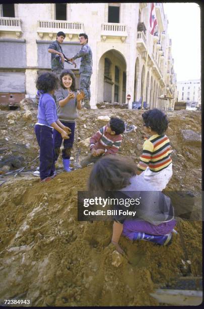 Moslem kids cavort atop dirt mound, digging with fingers, under watchful eye of Lebanese Army troops.