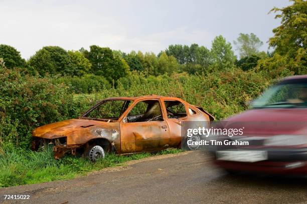 Car passes a burnt-out wreck by the roadside, Oxfordshire, United Kingdom.