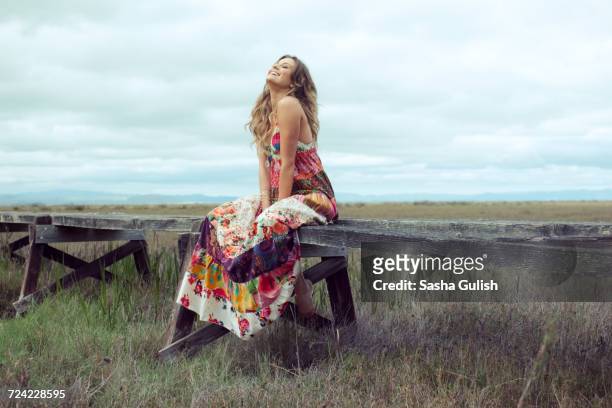 young woman in boho maxi dress sitting on elevated wooden walkway in landscape - long dress stock-fotos und bilder