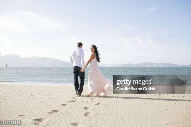 woman looking back while strolling hand in hand with boyfriend on beach - woman long dress beach stock pictures, royalty-free photos & images